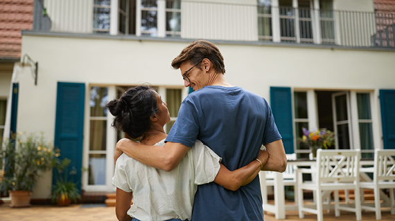 Zwei Personen stehen Arm in Arm vor einem Haus mit blauen Fensterläden und einer Terrasse mit weißen Gartenmöbeln.